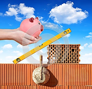 Masonry tools on a brick wall and a hand holding a piggy bank