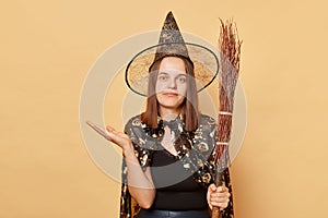 Masquerade party theme. October's magical holiday. Uncertain young woman wizard wearing witch costume holding in hand broom
