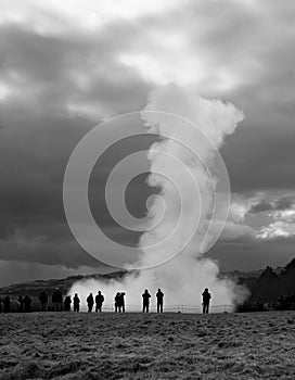 A massive geysir erupting among small shapes of tourists in wonder