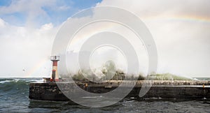 Massive wave crashing against a lighthouse during a storm with a rainbow in the sky.