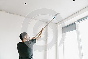 The master in a gray T-shirt paints the ceiling with a roller in the gray color in his apartment.