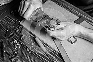 Master holding a hole punch and a piece of leather. On brown wooden table scattered with tools and accessories