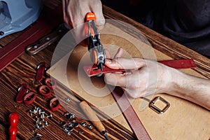 Master holding a hole punch and a piece of leather. On brown wooden table scattered with tools and accessories