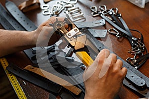 Master holding a hole punch and a piece of leather. On brown wooden table scattered with tools and accessories