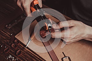 Master holding a hole punch and a piece of leather. On brown wooden table scattered with tools and accessories