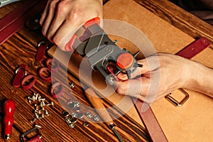 Master holding a hole punch and a piece of leather. On brown wooden table scattered with tools and accessories