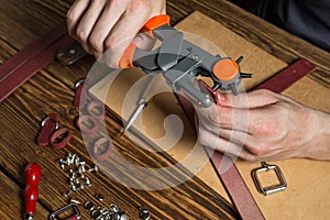 Master holding a hole punch and a piece of leather. On brown wooden table scattered with tools and accessories.