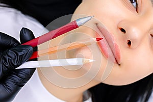 Master of permanent makeup in a black glove holds marking pencils in front of the model`s lips