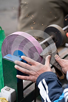 Master sharpens ax on electric sharpener with bright sparks and holds blade with his brutish fingers