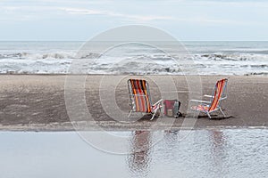 Mate set bag, yerba mate container, mate and thermos next to two beach chairs on the beach, reflected in the water.