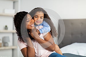 Maternal love. Cheerful black girl hugging her mommy on bed at home