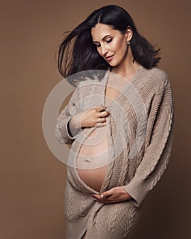 Maternity Knitwear. Pregnant Woman in Woolen Cardigan looking at Belly over Beige Background. Pregnancy Fashion Clothes. Maternal