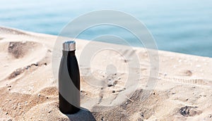 Matte black steel thermo bottle for water in sand of the beach on background of sea. Empty blank for mockup. Say no to plastic.