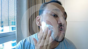 Mature adult asian metrosexual man applying shaving cream on face skin. Handsome bearded ethnic hipster guy puts facial creme at