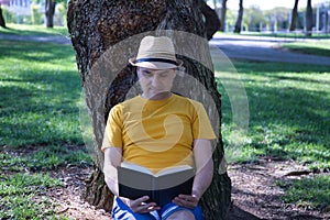 Mature adult Hispanic male sitting in a park, leaning against a tree, reading a book peacefully