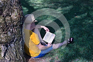 Mature adult Hispanic male sitting in a park, leaning against a tree, reading a book peacefully