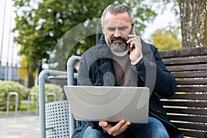 mature adult male lawyer with a beard works using a laptop and speaks on the phone on the street