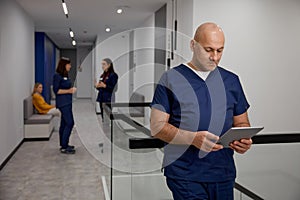 Mature adult man doctor in uniform with digital tablet in clinic corridor