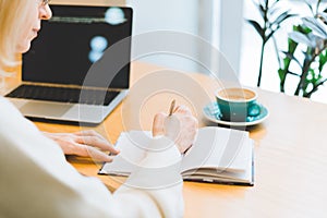 Mature adult woman sitting in cafe with coffee mug and working online on laptop computer. Businesswoman in eyeglasses