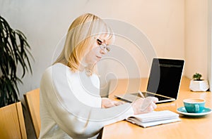 Mature adult woman sitting in cafe with coffee mug and working online on laptop computer. Businesswoman in eyeglasses