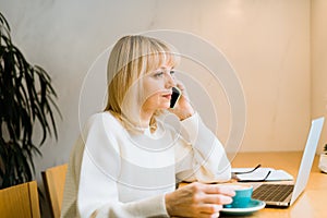 Mature adult woman sitting in cafe with coffee mug and working online on laptop computer. Businesswoman talking over the