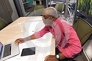 Mature african american engineer using laptop while sitting with blueprint at desk in workplace