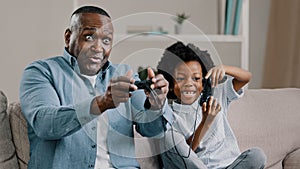 Mature african american father with little daughter playing video games on console use joystick controller sitting on