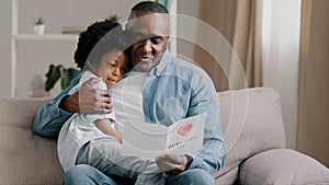 Mature african american man with cute kid girl hugging sitting in room on sofa father reads greeting card loving