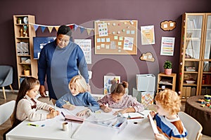 Mature African American nursery teacher standing by table where kids drawing