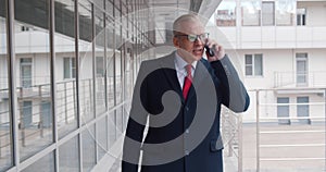 Mature angry businessman talking on mobile phone walking outside business center in city