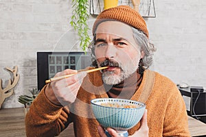 Mature Asian man with a white beard and long hair eating noodle dish with chopsticks