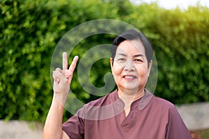 Mature asian woman standing and showing 2 fingers at public park,Happy and smiling,Senior care insurance concept