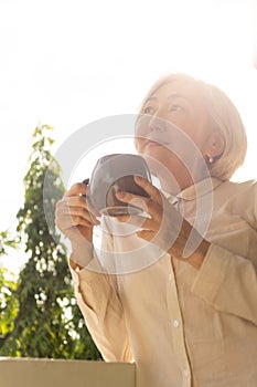 Mature asian woman in sun light on balcony with cup of coffee. Morning, lifestyle concept, text space