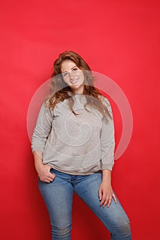 Mature attractive brunette woman in grey sweater and blue jeans standing on red studio wall background