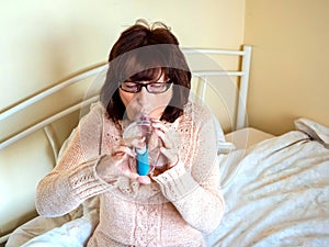 Mature attractive lady sitting on her bed using asthmatic pump spacer device to ease condition