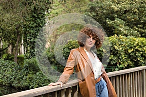 Mature and attractive woman, with curly brown hair, wearing a brown leather jacket, shirt and jeans, leaning on a wooden railing