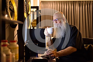 Mature bald bearded man drinking coffee while working overtime at home late at night