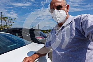 A mature bald Caucasian man wearing a white face mask with his arm on the trunk of a white car photo