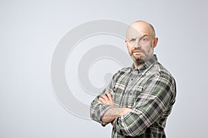 Mature bald man in checkered shirt standing with arms crossed and serious face at camera, looking suspicious