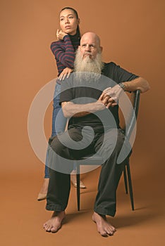 Mature bearded bald man with young Asian transgender woman against brown background