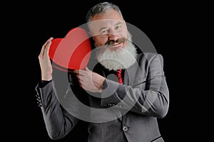 Mature bearded man wear suit holds red heart shaped gift box  isolated black background