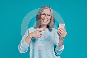 Mature beautiful woman pointing smart phone and smiling while standing against blue background