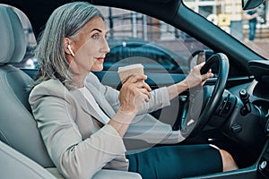 Mature beautiful woman in smart casual wear smiling and drinking coffee while driving car