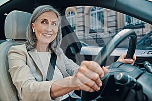 Mature beautiful woman in smart casual wear smiling and driving while sitting on front seat of the car