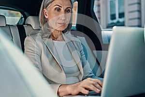 Mature beautiful woman in smart casual wear using laptop while sitting on the back car seat