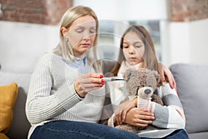 Mature blond woman, checking her sick daughter's temperature on thermometer after measuring. Sad ill girl, holding teddy