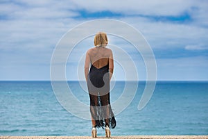 Mature, blonde, beautiful woman wearing an elegant black dress gazes longingly at the horizon over the atlantic ocean in cadiz, photo