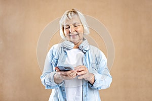 Mature blonde woman with smart phone posing isolated on beige background, studio portrait. Cute beautiful 60s woman with