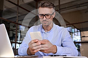 Mature business man holding cell using phone checking data working in office.