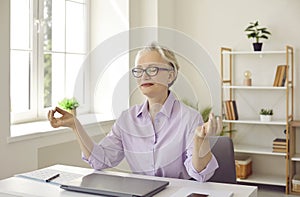 Mature business woman meditates at office working desk, relieves stress, feels calm and peaceful